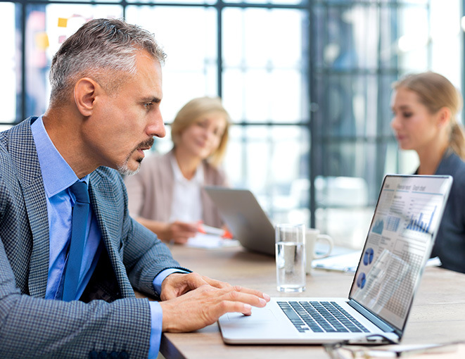 A man in a suit working intently on a laptop, analyzing charts and graphs displayed on the screen. In the background, two colleagues are visible, seated at a conference table in a modern office with large windows. The image represents the concept of Executive Search, highlighting detailed analysis, strategic recruitment, and a collaborative professional environment.